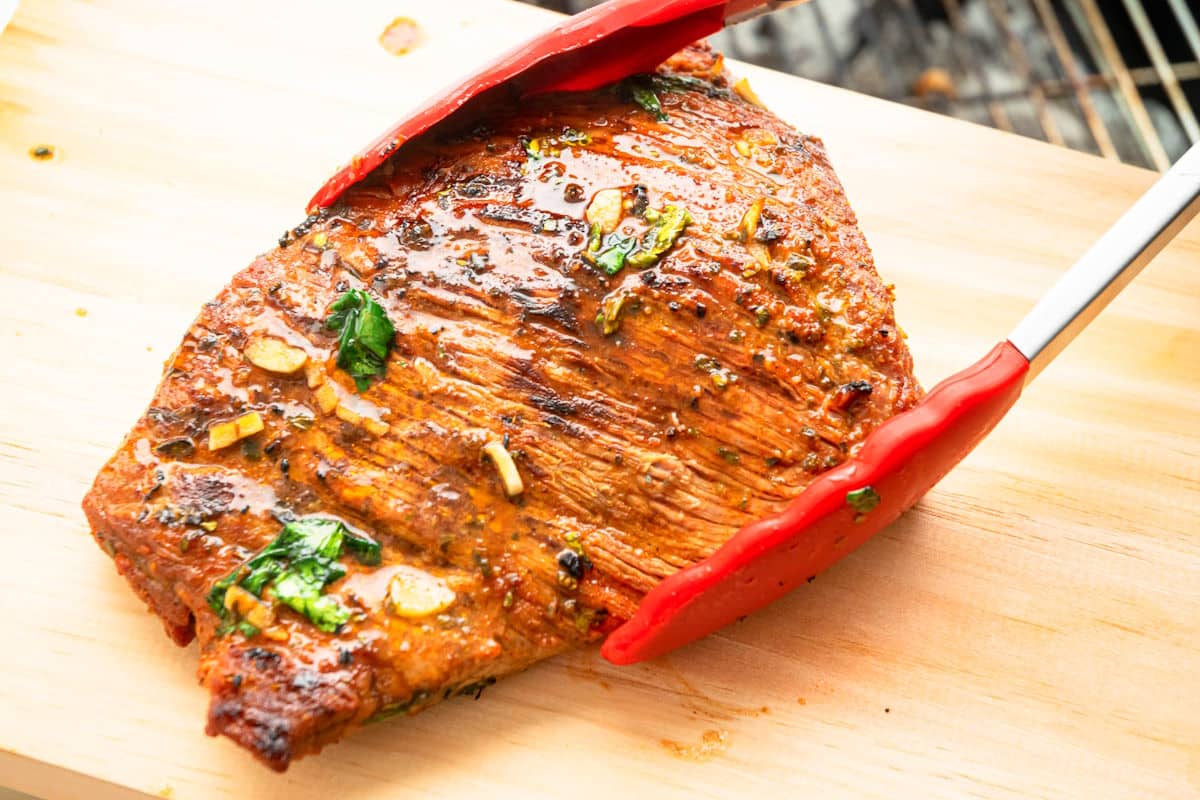 Grilled carne asada on cutting board in tongs.