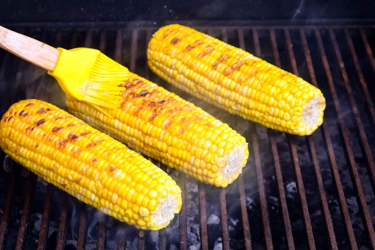 Grilled corn on the cob on grill grates getting brushed with butter.