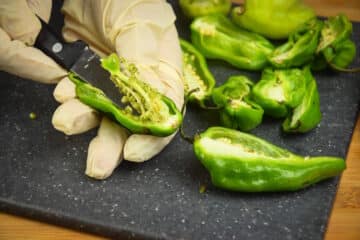 Jalapeño peppers sliced open on cutting board and gloved hand holding paring knife.