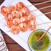 Jumbo shrimp on skewers on white dish with chimichurri on the side.