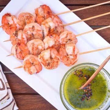 Jumbo shrimp on skewers on white dish with chimichurri on the side.