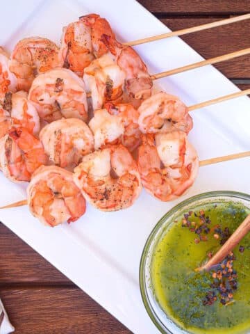 Jumbo shrimp on skewers on white dish with chimichurri on the side.
