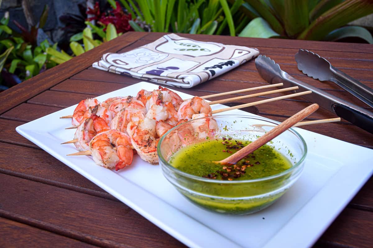 Grilled jumbo shrimp skewers on white dish with bowl of chimichurri sauce.