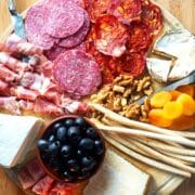 Circular charcuterie board with meat, cheese, breadsticks, nuts, apricots and olives.