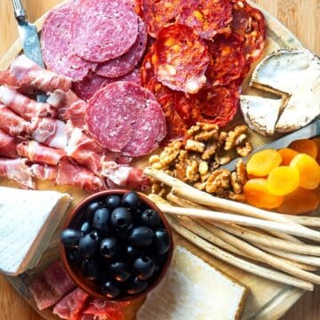 Circular charcuterie board with meat, cheese, breadsticks, nuts, apricots and olives.