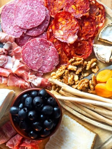 Circular charcuterie board with meat, cheese, breadsticks, nuts, apricots and olives.