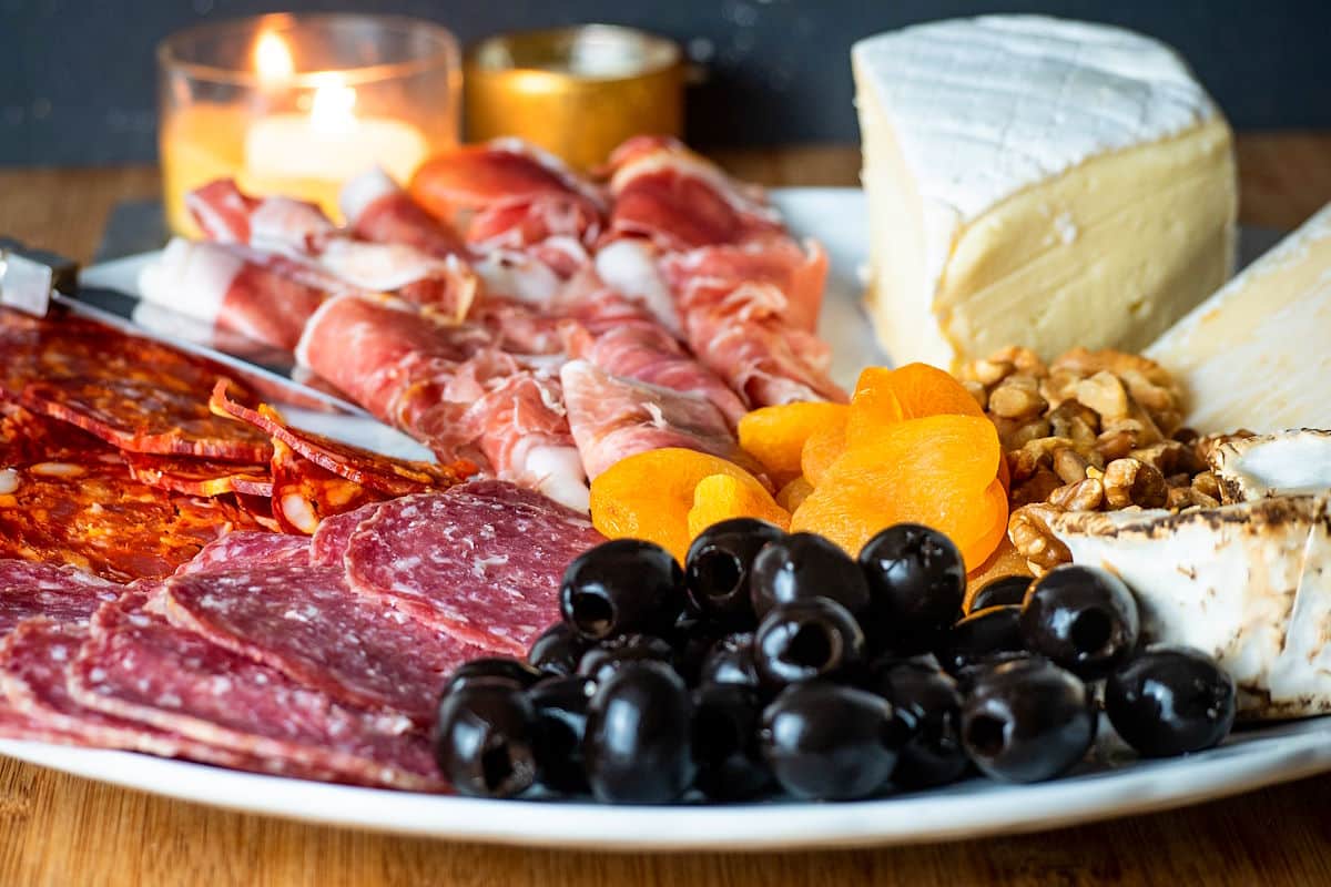 Circular charcuterie board with meat, cheese, breadsticks, nuts, apricots and olives.