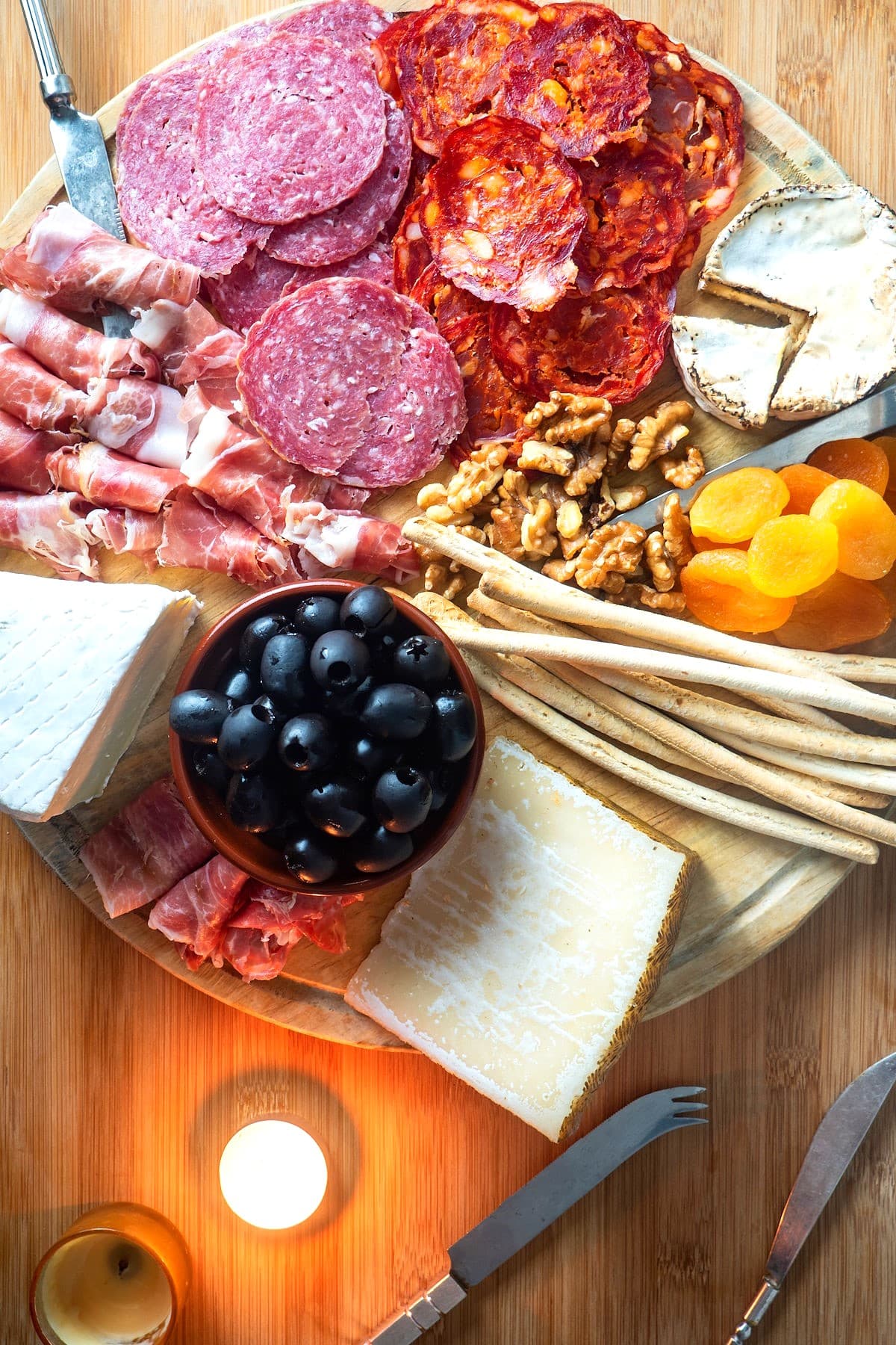 Circular charcuterie board with meat, cheese, breadsticks, nuts, apricots and olives.