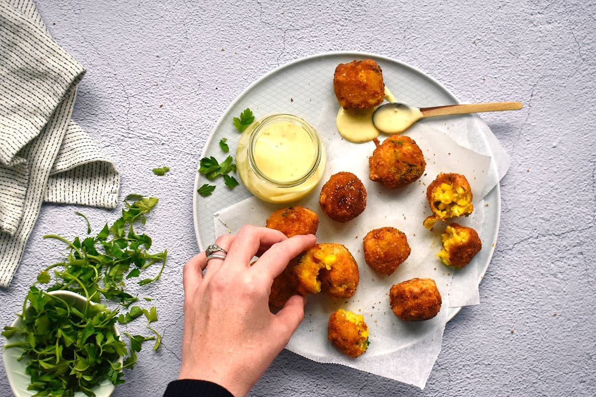 A woman's hand picking up fried corn nuggets on plate with dip.