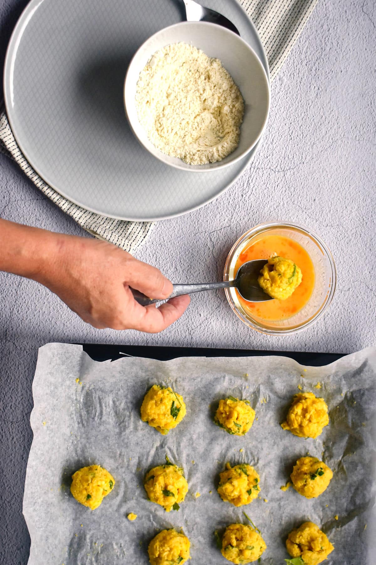 Raw corn nuggets on baking sheet with wax paper and dipped into egg mixture on spoon.
