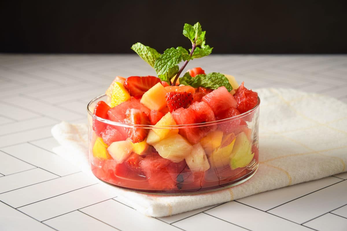 Fruit salad in a glass dish on white tile and black background.