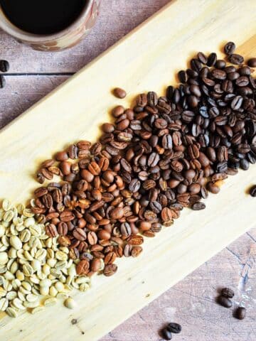 4 colors of coffee beans on wooden board with a mug of coffee on the side.