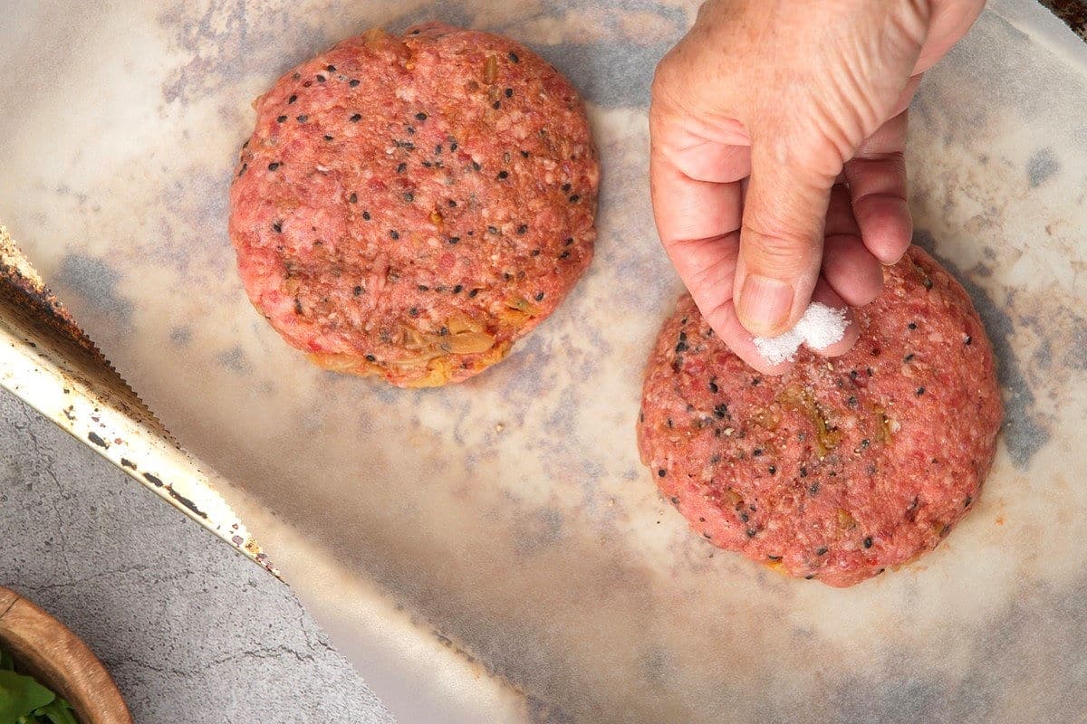 Kimchi beef patties on parchment lined baking sheet and woman's hand sprinkling them with salt.