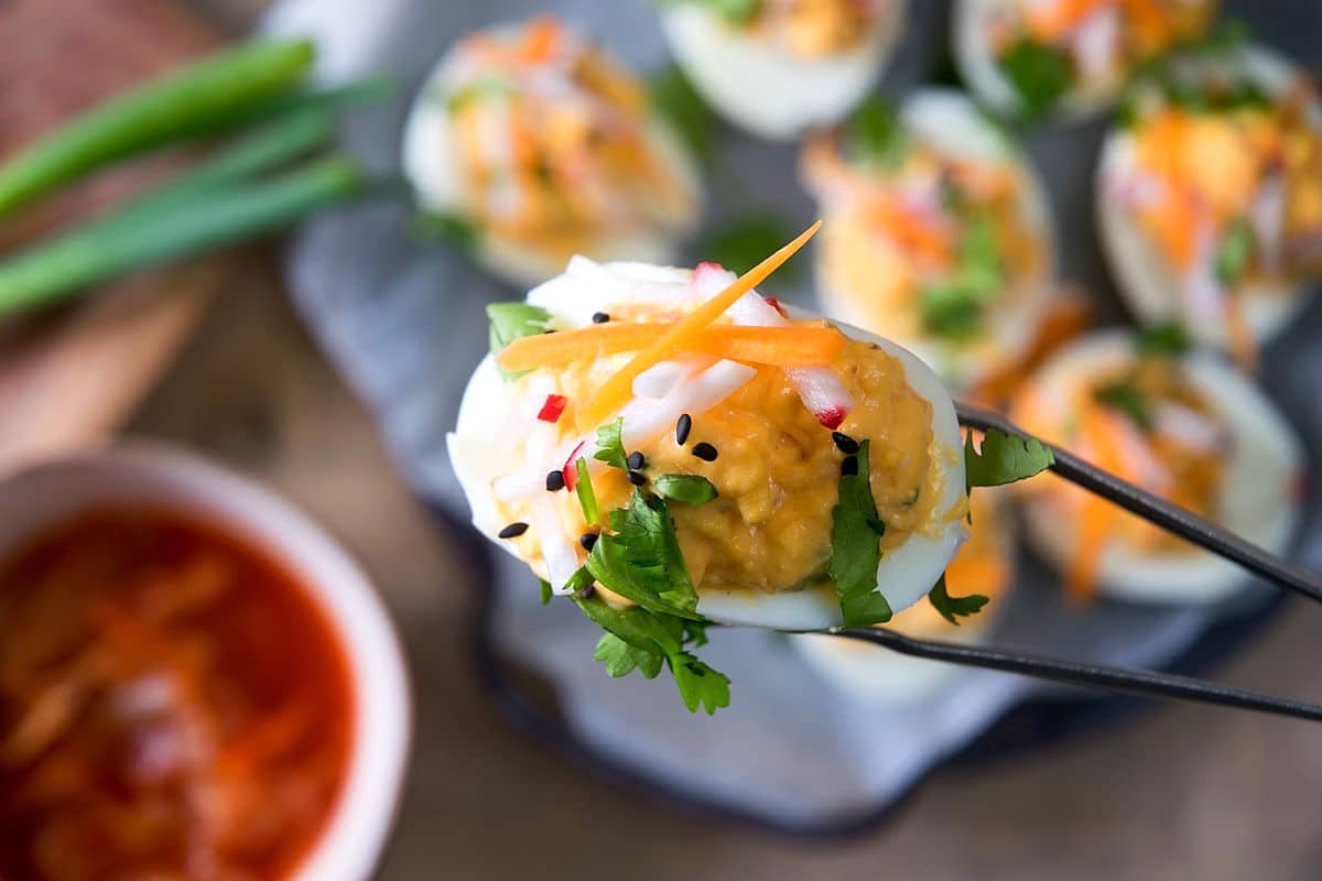 Deviled egg with kimchi and pickled veggies held by cooking tweezers.