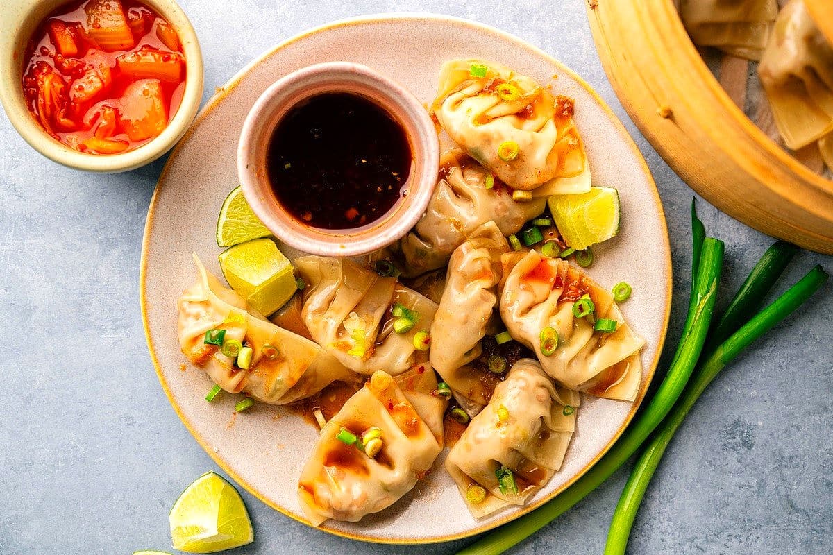 Kimchi dumplings arranged on plate with dipping sauce, small bowl of kimchi on the side.
