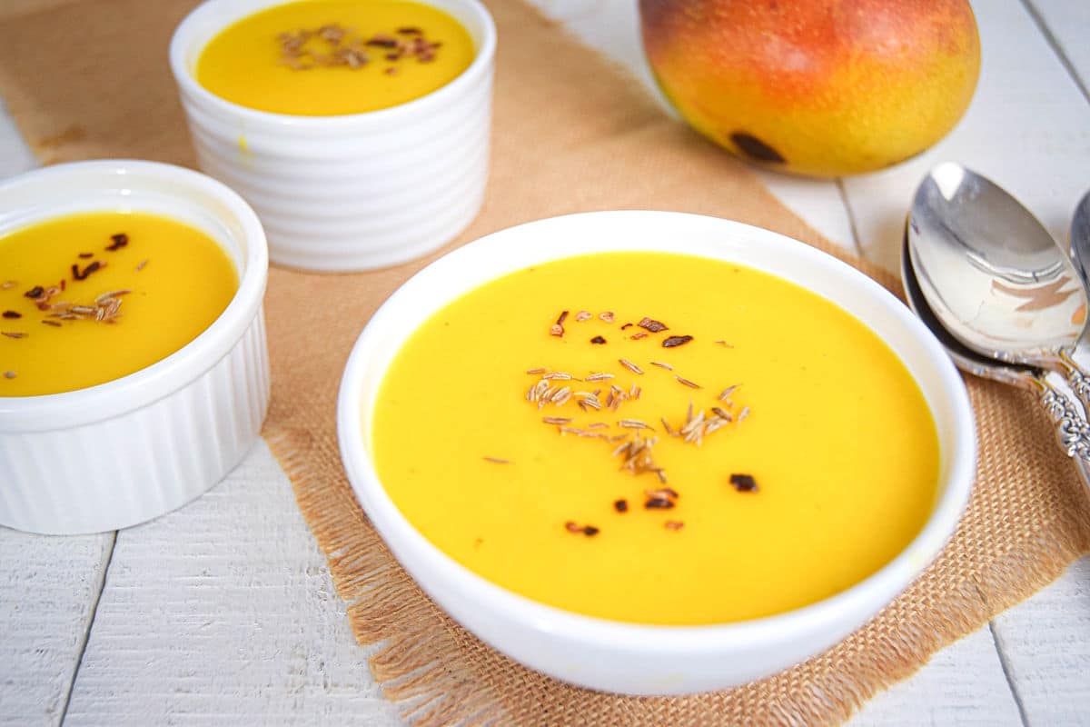Mango soup in white bowls with a fresh mango and spoons on the side.