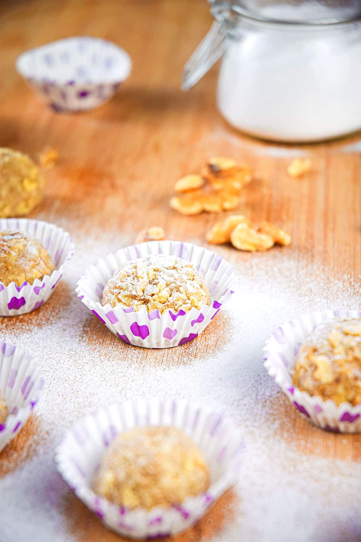 Marzipan balls dusted in icing sugar in white and purple baking cups with nuts on the side.
