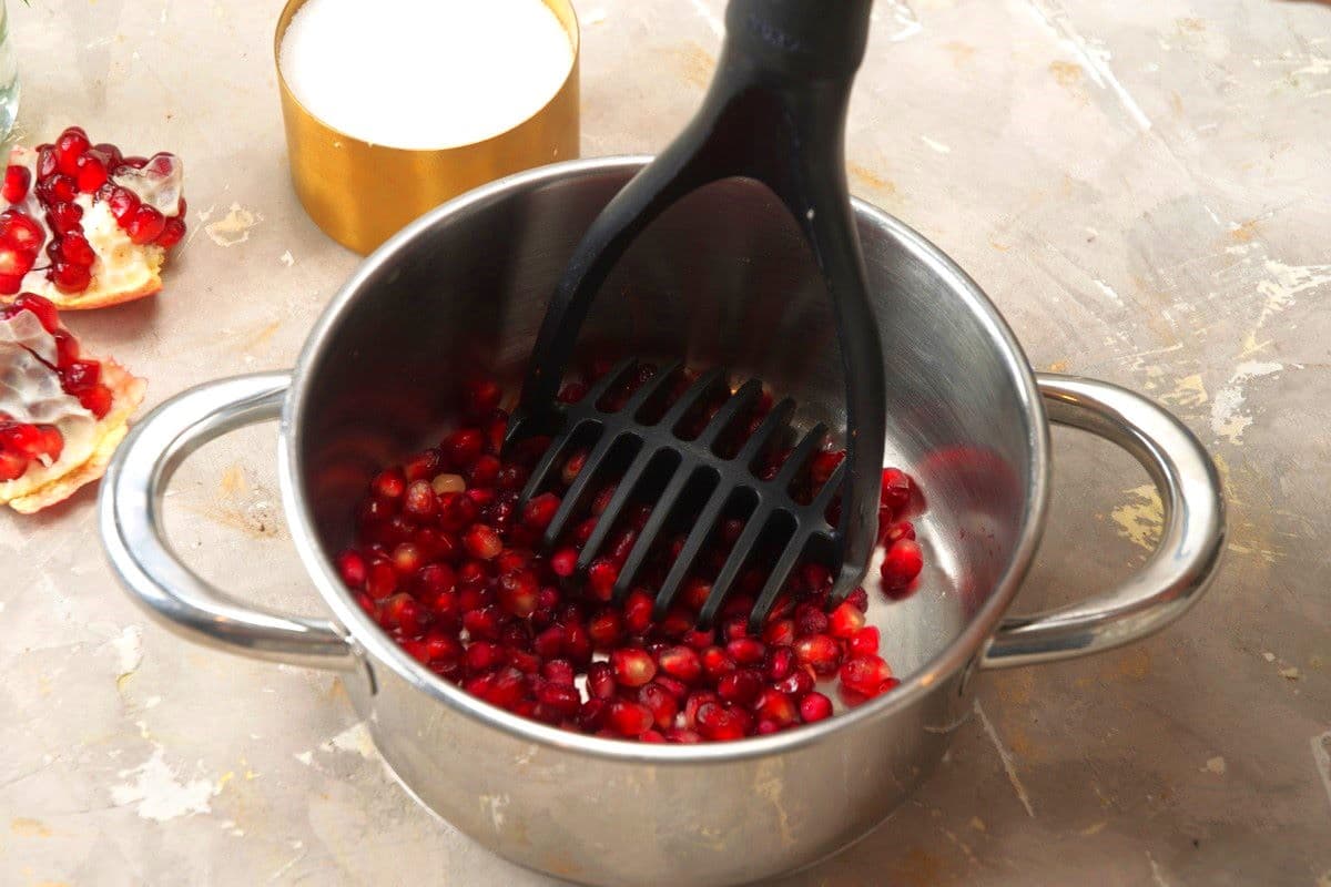 Pomegranate seeds in saucepan with potato masher.