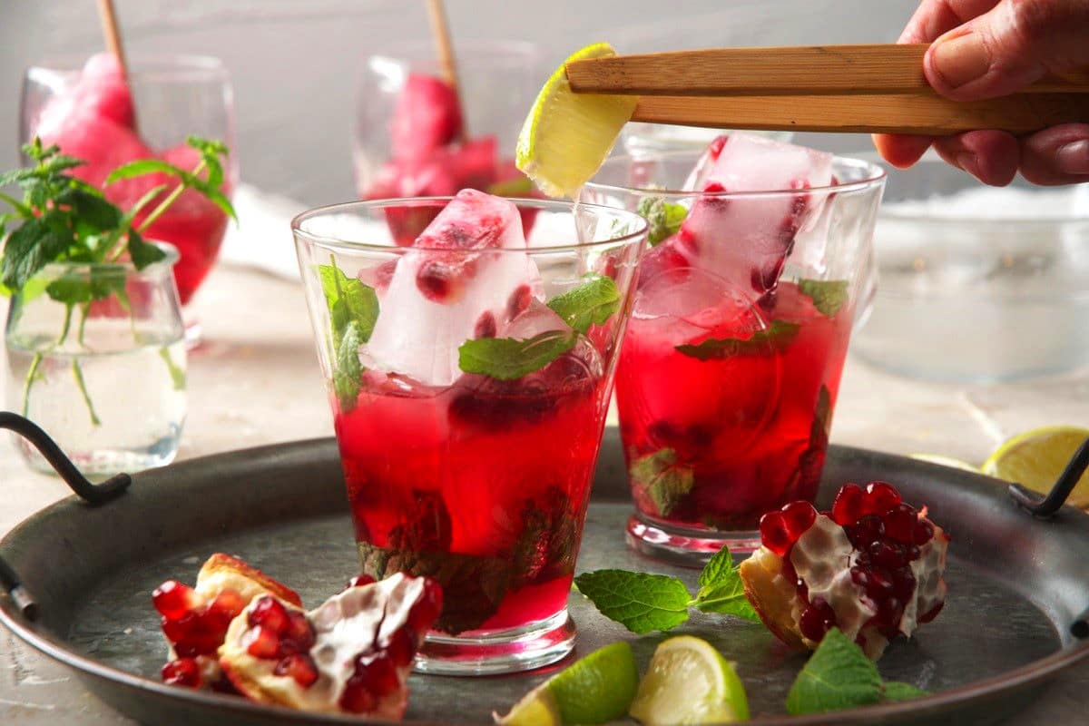 Pomegranate seeds, mint and ice cubes in glasses with lime wedge in cooking tweezers.