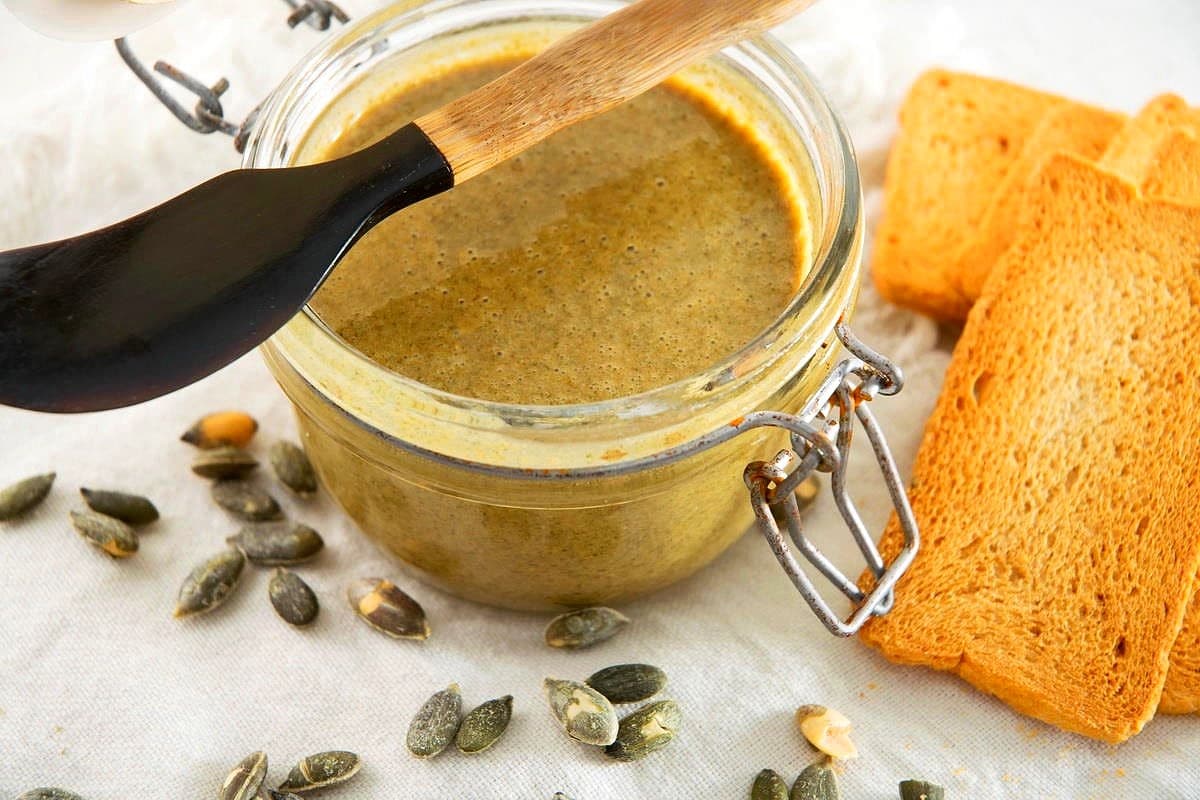 Pumpkin seed butter in jar with mini spatula on top and pumpkin seeds on the side.