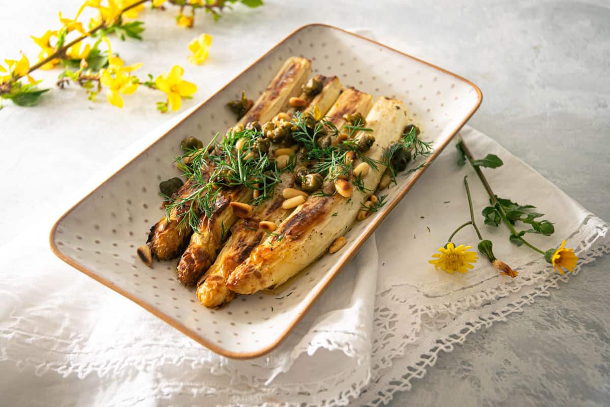 Roasted white asparagus on white dish with capers and dill and yellow flowers on the side.