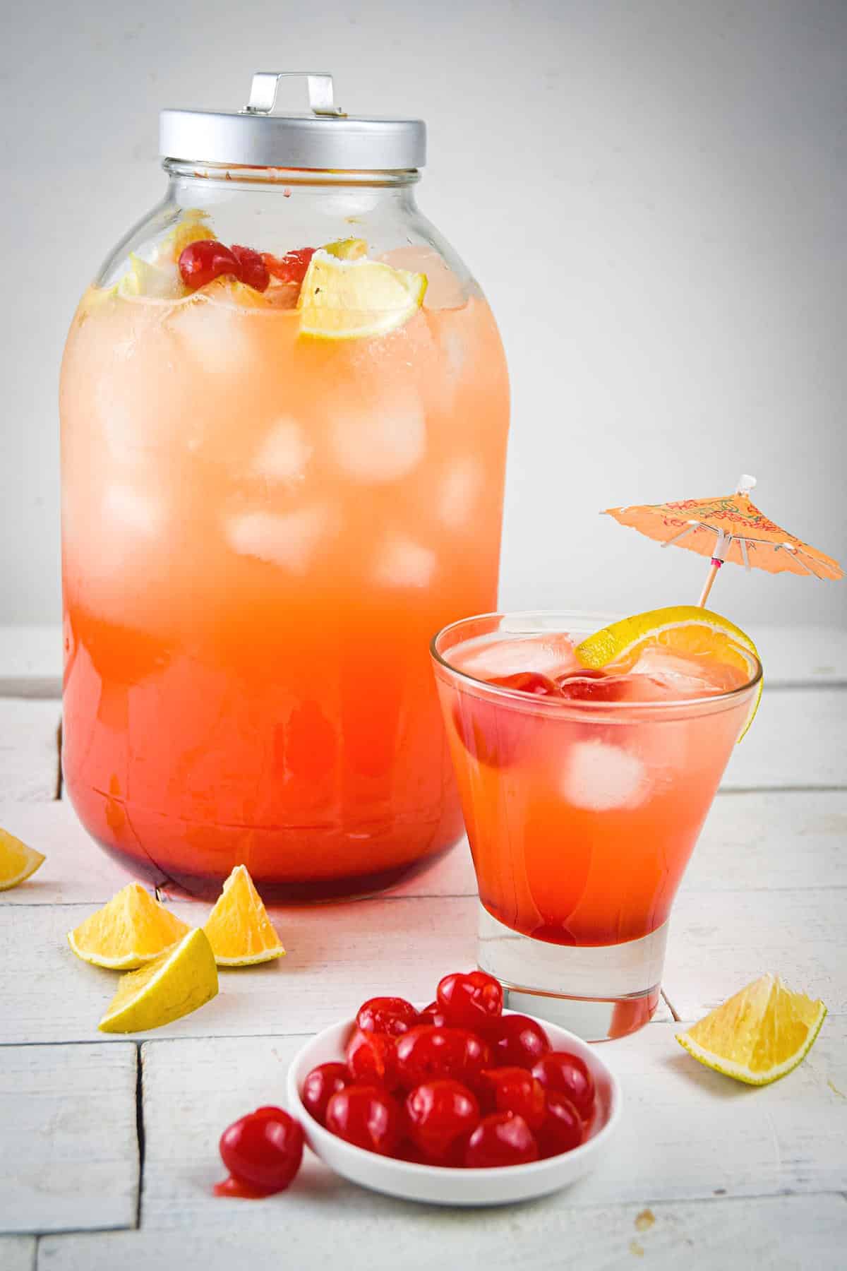 Shirley temple punch in glass and pitcher with maraschino cherries and orange wedges on the side.