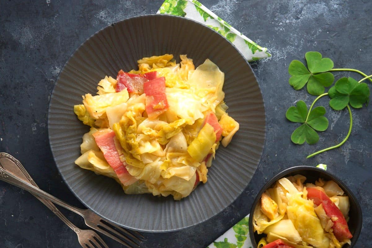 Cooked cabbage and bacon in black bowls and 4 leaf clovers on the side.