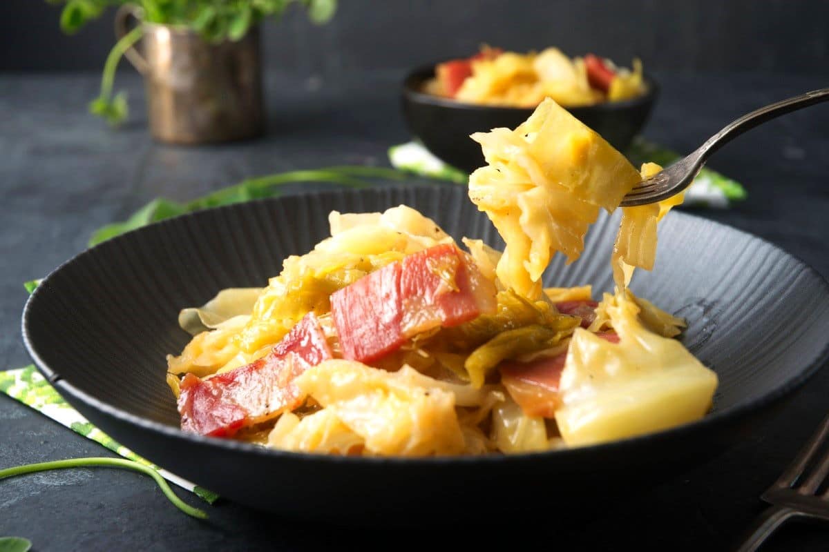 Cooked cabbage and bacon in black bowl with fork.