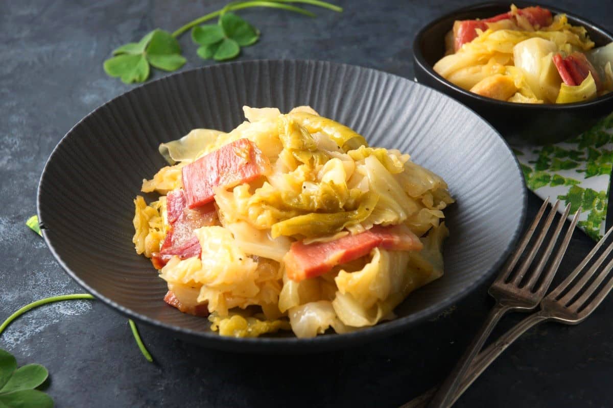 Cooked cabbage and bacon in black bowl with forks on the side.