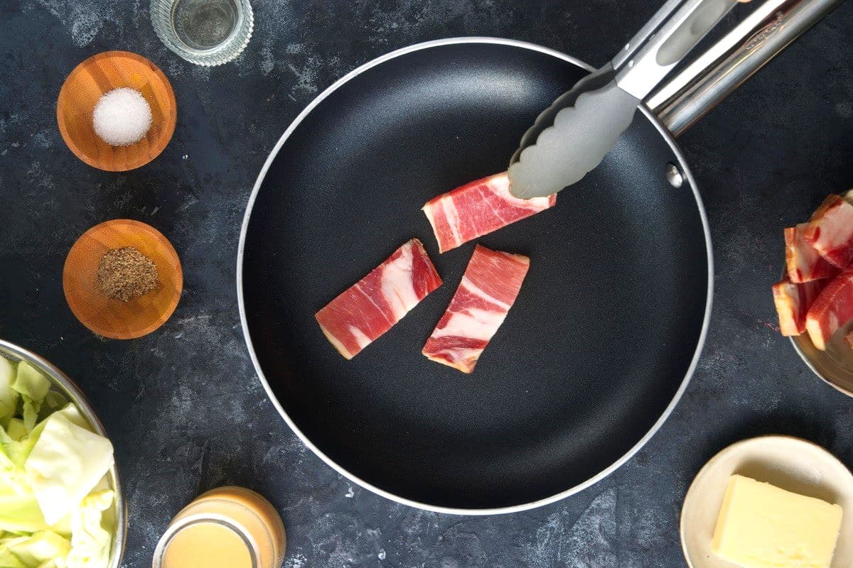 Raw bacon strips in pan with tongs.