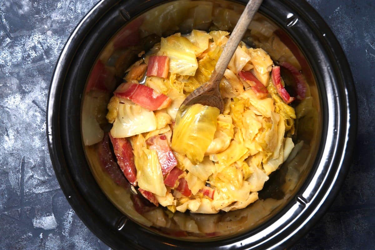 Cabbage and bacon in slow cooker with wooden spoon.