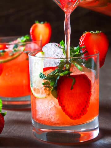 Strawberry mojito mocktail pouring out of pitcher into glass with ice cubes.