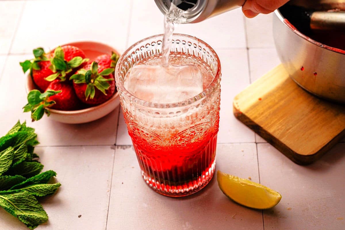 Sparkling water pouring into glass with strawberry syrup, lime, ice and mint.