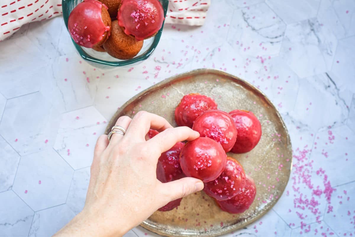 A woman's hand picking up a Valentine's day cake ball with pink frosting and sprinkles.