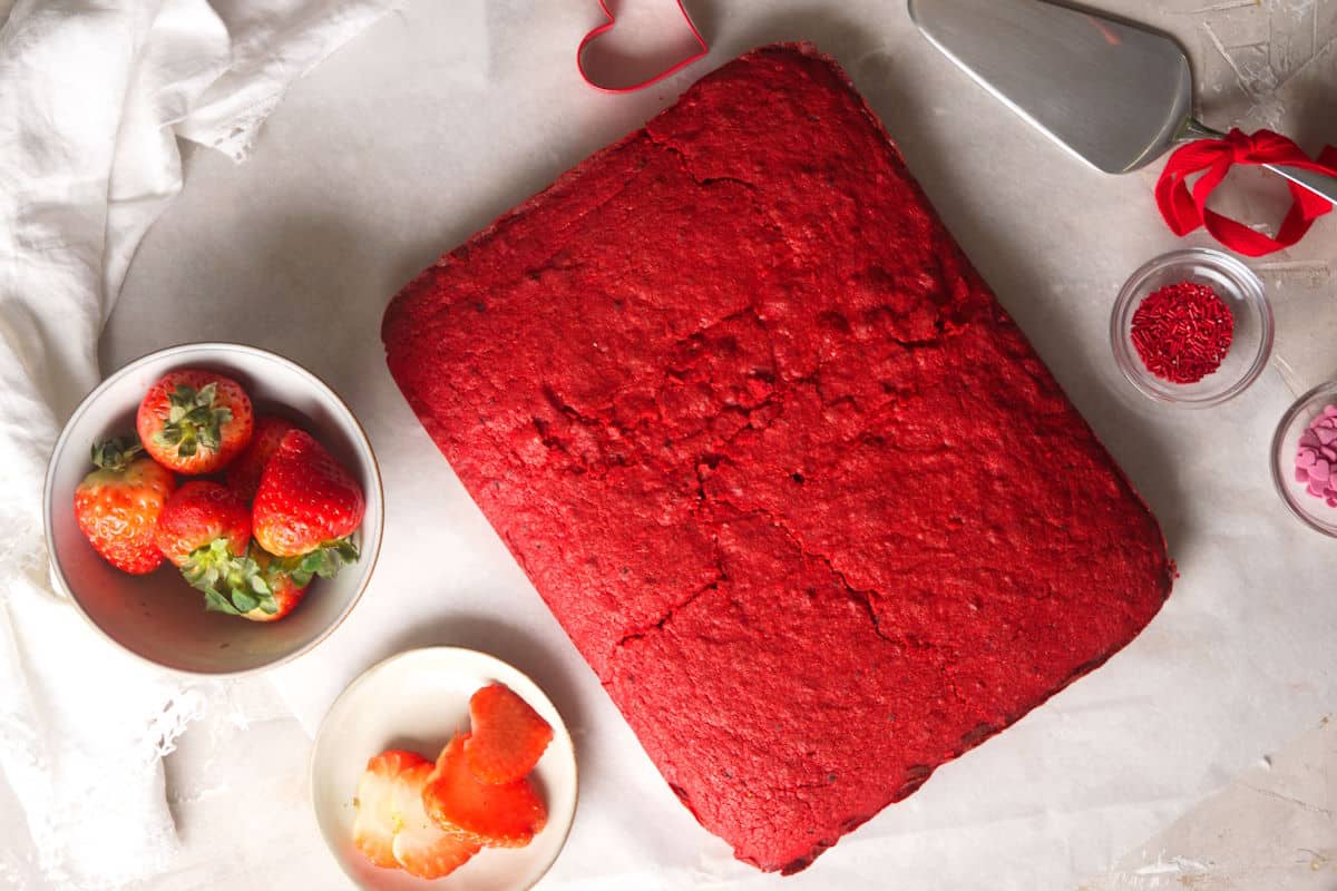 Red velvet sheet cake on parchment with strawberries and candy decorations on the side.