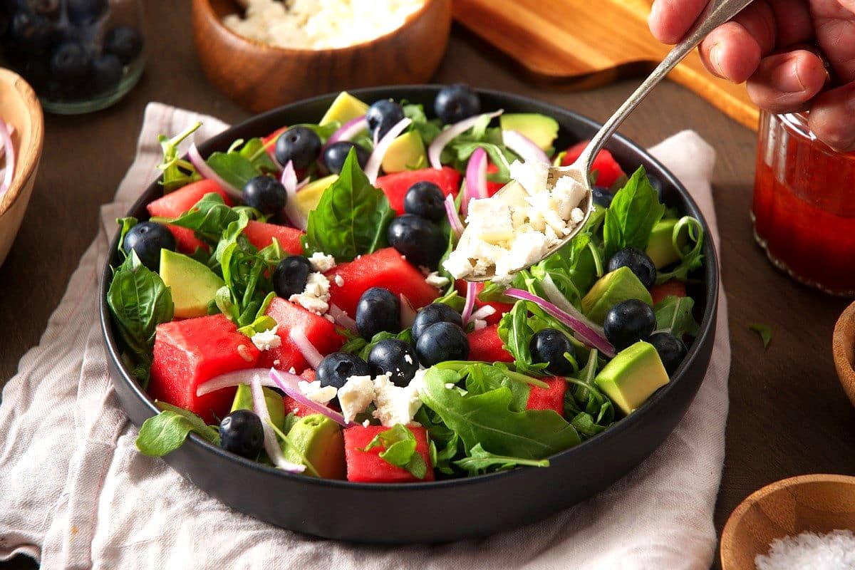 Watermelon salad in black bowl with feta getting sprinkled on top.